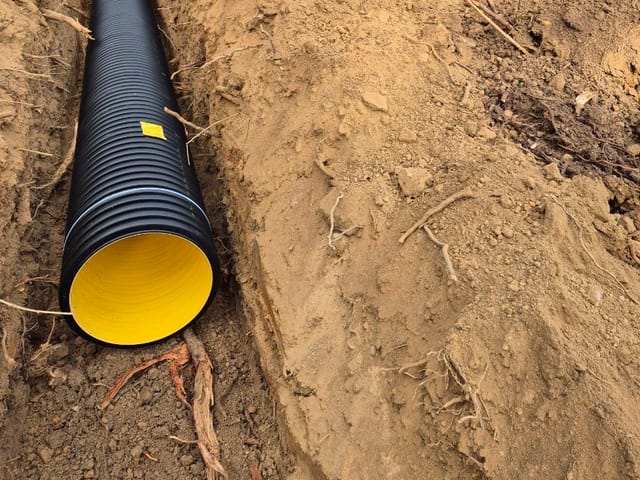 Large black corrugated drainage pipe laid in a freshly dug trench, with the spoil heaped alongside