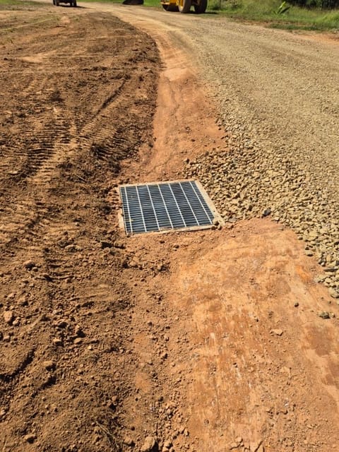New steel grate drainage pit set into graded ground beside a gravel driveway