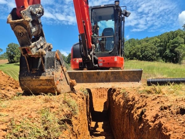 Orange Kubota excavator at the end of a deep, clean-walled trench in red soil, with drainage pipe ready beside it