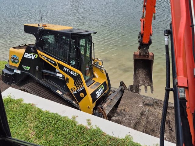 ASV RT-60 posi-track and excavator digging along a canal edge beside a concrete edging strip